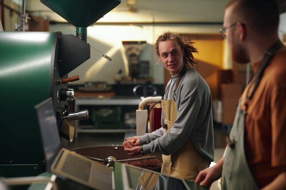 Male coffee roaster chatting with his colleague while they controlling process of roasting coffee beans.
